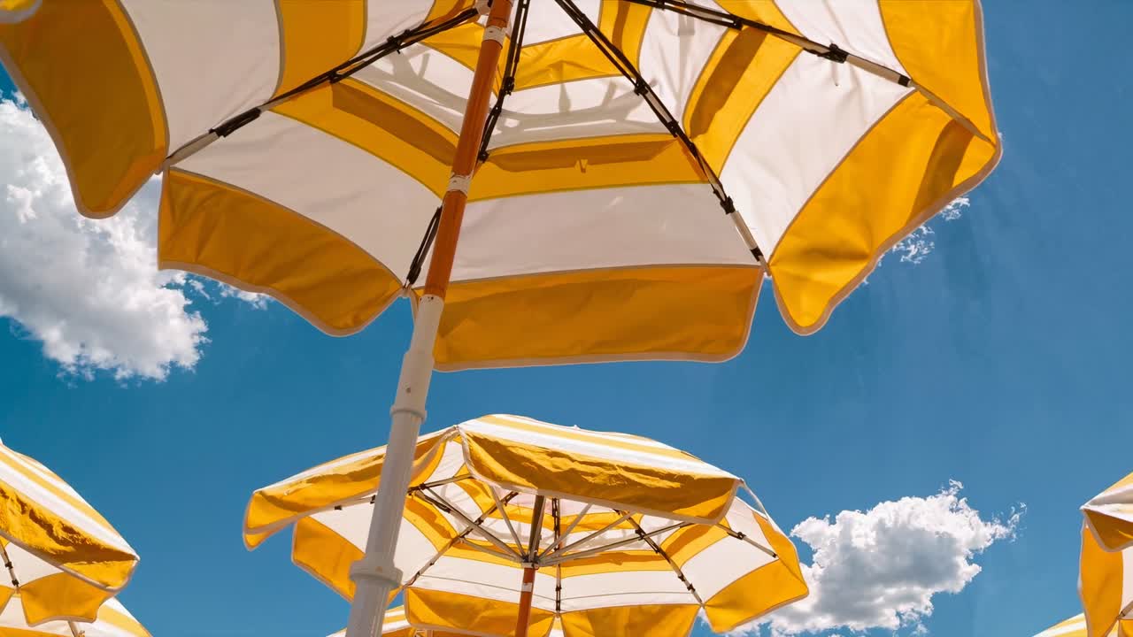 Ripresa video dal basso di ombrelloni da spiaggia gialli e bianchi contro un cielo blu brillante con soffici nuvole.