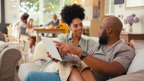 Couple-At-Home-Using-Laptop-To-Shop-Or-Book-Holiday-With-Multi-Generation-Family-In-Background