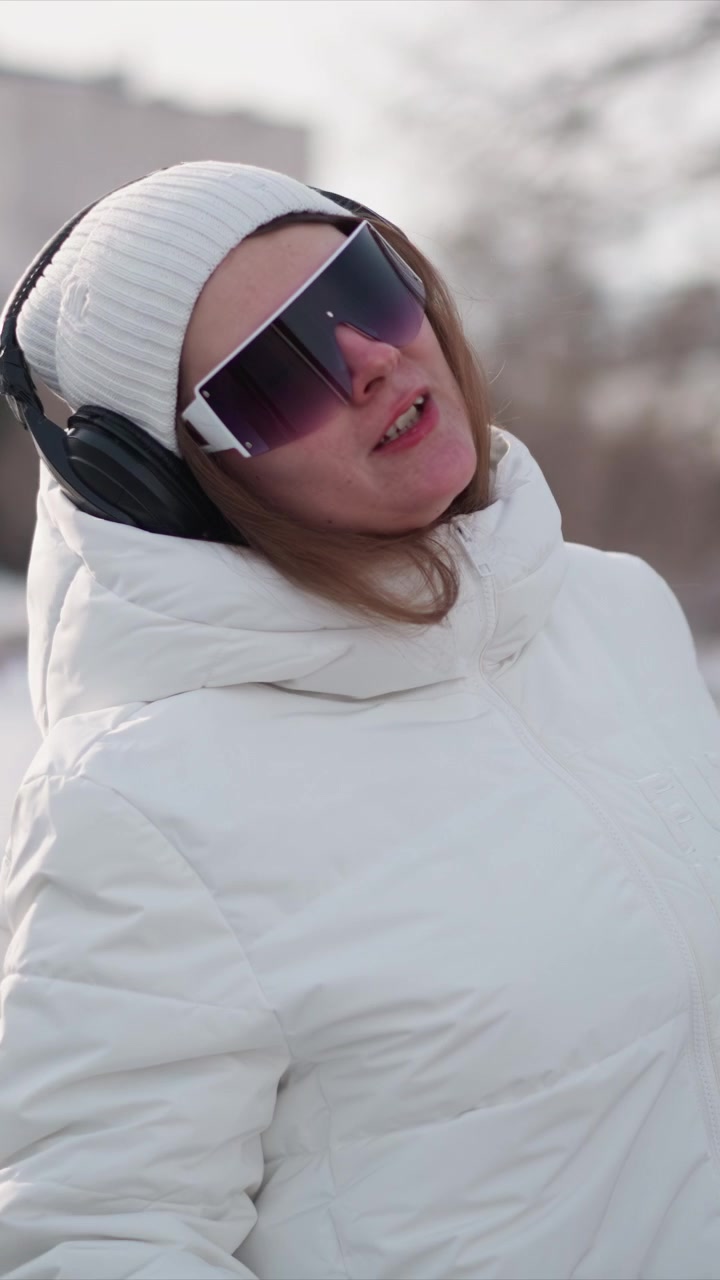 Woman wearing white puffer jacket black headphones and sunglasses moves through outdoor winter environment. Her hair flows as woman turns her head or walks with background showing blurred
