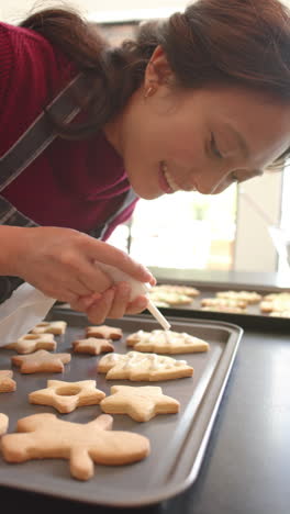vertical video of happy biracial woman decorating chistmas cookies in kitchen, slow motion