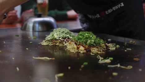 chef prepares hiroshima style okonomiyaki with noodles on large teppan grill