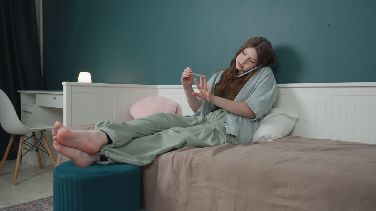 Ragazza adolescente bianca che si smalta le unghie sul letto mentre è al telefono, si rilassa in comodo abbigliamento da casa, luce serale soffusa, ottomana e cuscino soffice visibili, manicure e conversazione multitasking, cura di sé casual.