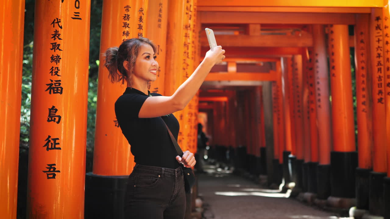 mujer con cabello marrón claro en cola de caballo, con una camiseta negra y vaqueros, toma una selfie en el santuario fushimi inari, kyoto vibrantes puertas de torii naranja con inscripciones japonesas profundidad de campo poco profunda