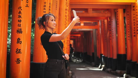 Woman-with-light-brown-hair-in-a-ponytail,-wearing-a-black-top-and-jeans,-takes-a-selfie-at-Fushimi-Inari-Shrine,-Kyoto-vibrant-orange-torii-gates-with-Japanese-inscriptions-shallow-depth-of-field