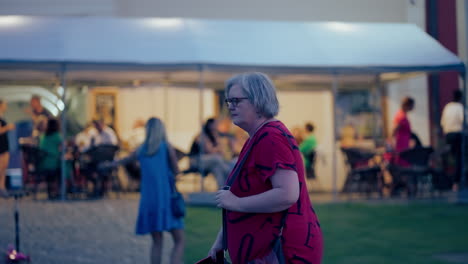 panning shot of a white woman with glasses wearing red t-shirt walking around historical city bardejov during a blue hour at night in slow motion, surrounded by people and lights