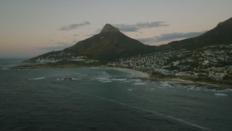 Toma-Panorámica-De-La-Bahía-Del-Mar-Con-Playa-De-Arena-Y-Edificios-En-El-Distrito-Urbano-Al-Atardecer.-Ciudad-Del-Cabo,-Sudáfrica