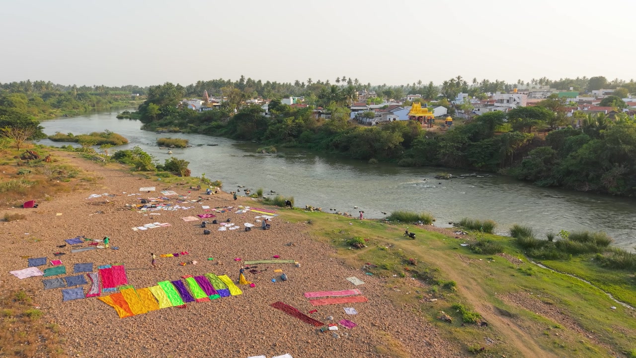 Aerial view of a riverside village where people dry colorful clothes and wash vehicles along the riverbank. Traditional rural laundry scene, calm flowing river, and peaceful countryside environment.