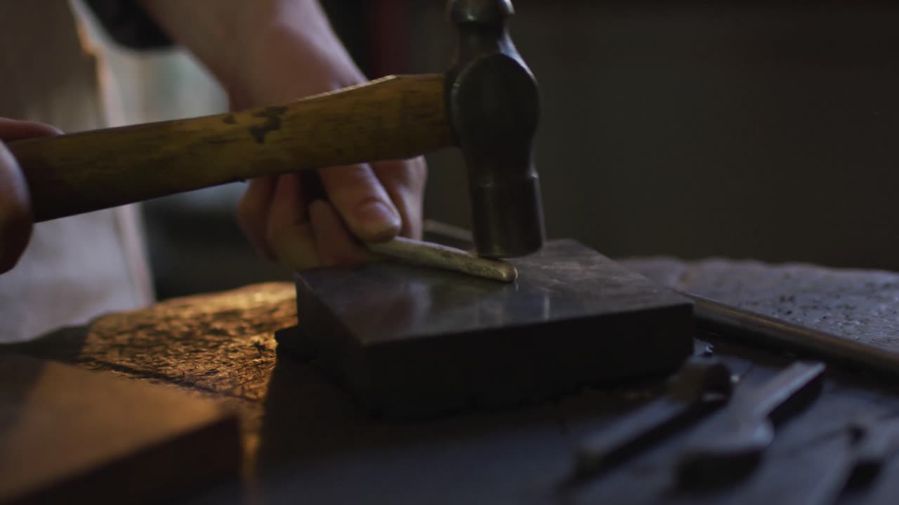 코카시안 여성 보석가의 손이 가까이 가서 망치를 사용하여 보석을 만들고 있습니다.
