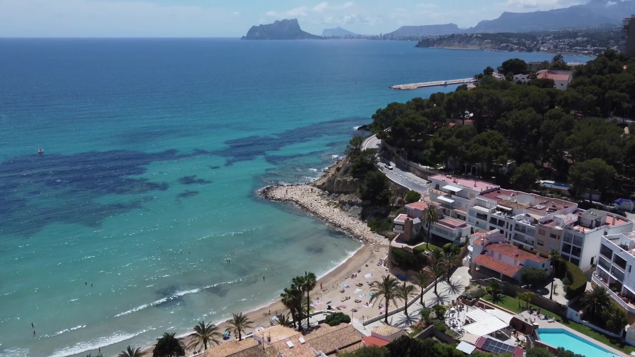Video aereo con drone che si inclina verso l'alto dalla spiaggia di El Portet a Moraira fino all'iconica cima del monte Peñón de Ifach a Calpe, Alicante, Spagna. Filmato stock della costa mediterranea turchese.