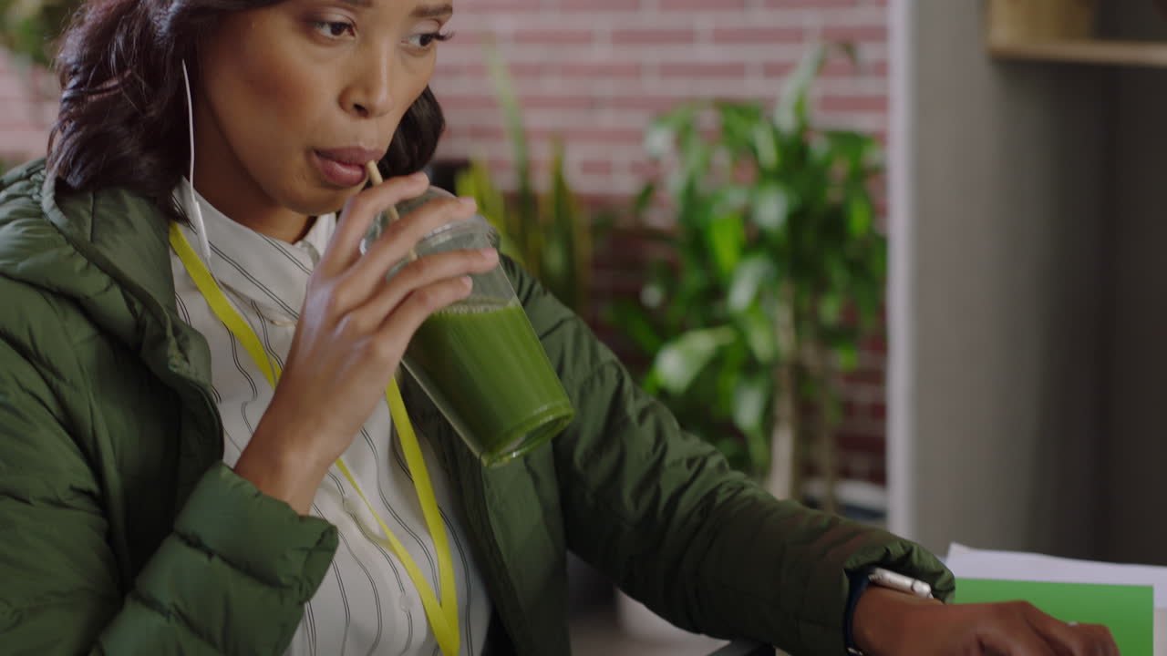 hermosa mujer de negocios afroamericana usando computadora portátil escribiendo navegando mensajes en línea bebiendo jugo escuchando música revisando teléfono inteligente en una oficina moderna y de moda