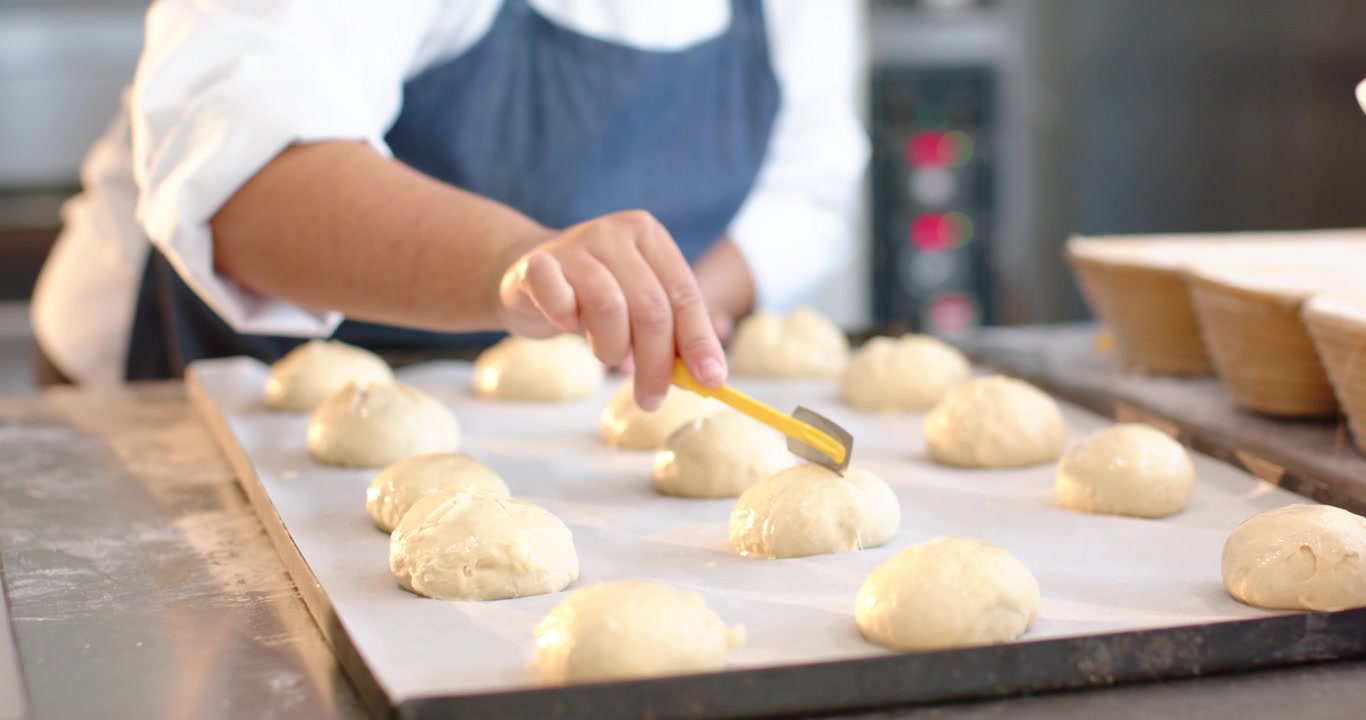 Adult woman baker scoring dough rounds with yellow lame on tray in bakery, preparing for baking