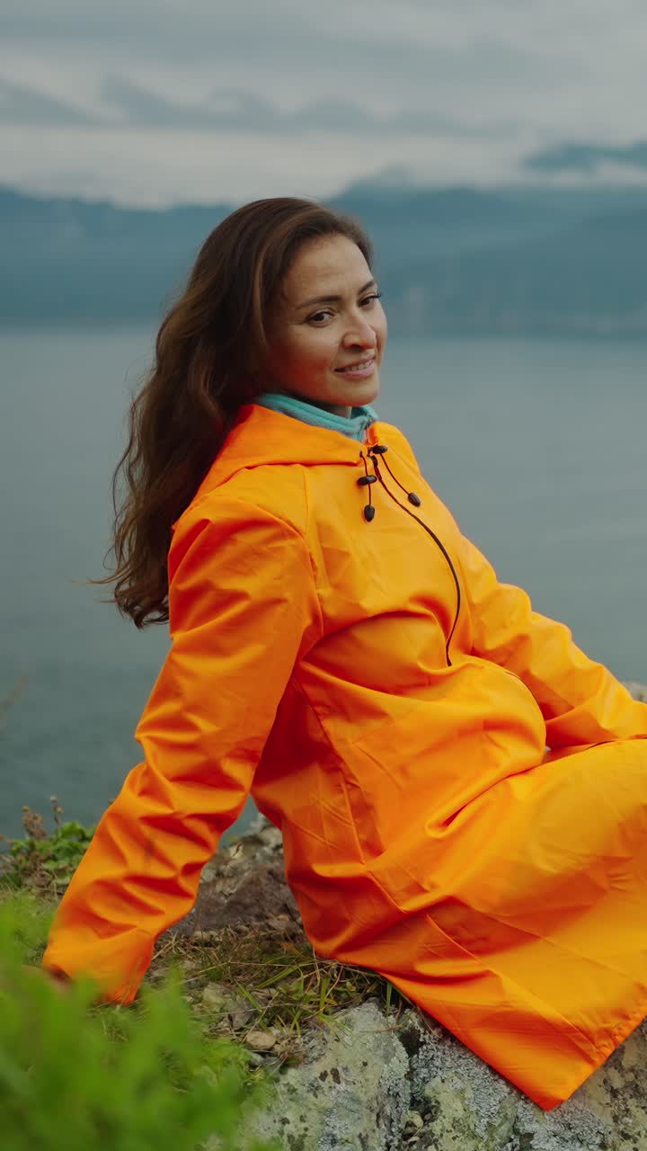 Donna con impermeabile arancione seduta in riva al lago e sorridente