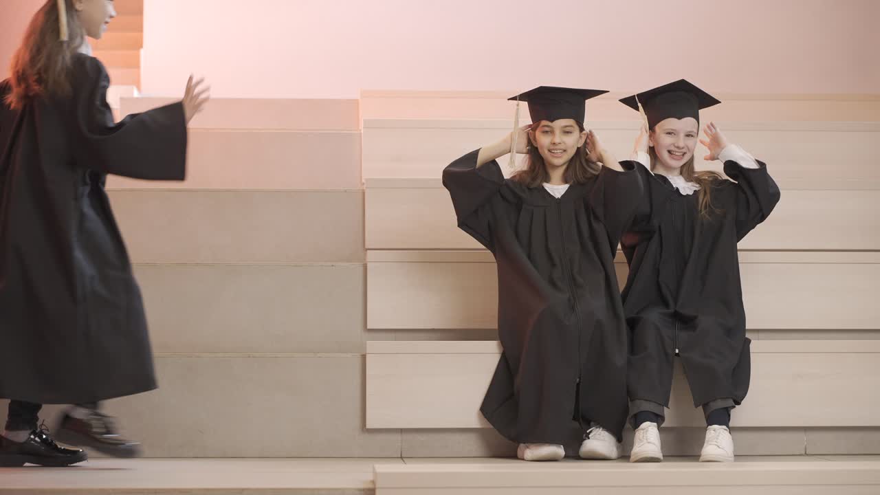 Three happy little girls in cap and gown playing together while sitting on stairs at the preschool graduation ceremony