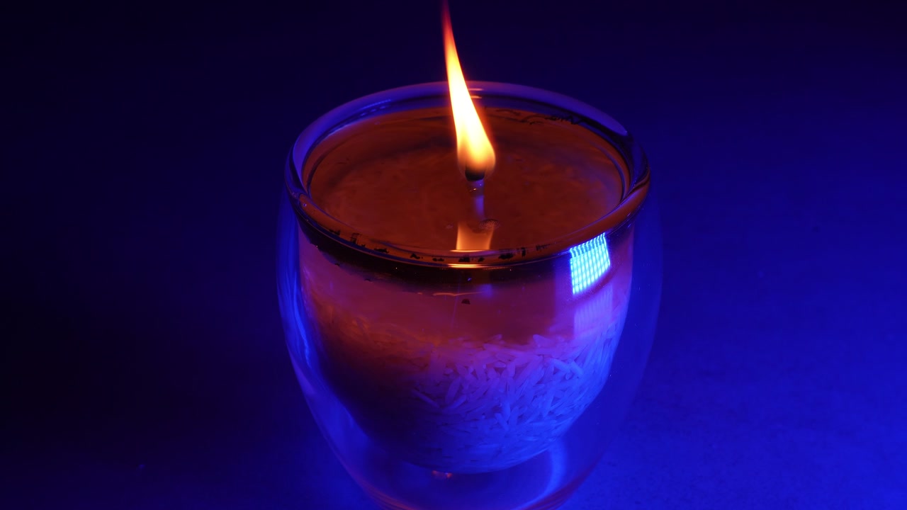 Un primo piano di una fiamma giallo brillante che brucia all'interno di un portacandele in vetro trasparente a doppia parete. L'ambiente dello studio interno è illuminato da una luce blu intensa, che si riflette nella pozza di cera liquida.