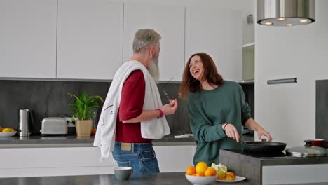 a happy elderly man with a beard in a red t-shirt and a brunette girl in a green sweater dance while preparing breakfast in the kitchen of a modern apartment