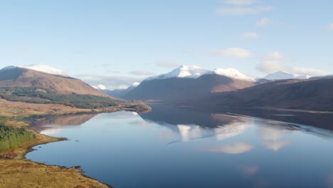 backwards flying aerial drone video revealing an epic winter view of glen etive and loch etive in the highlands of scotland with snow-capped mountains, a forest and beautiful still reflective water