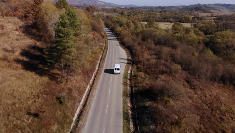 Nach-Einem-Weißen-Lieferwagen,-Der-An-Einem-Sonnigen-Tag-Auf-Einer-Herbstfarbenstraße-Fährt---Drohnenaufnahme-Aus-Der-Luft