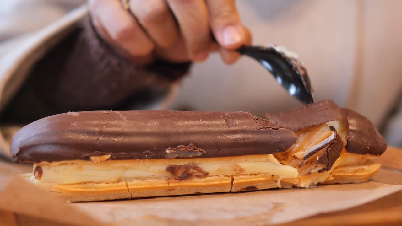 Close-up de uma mão comendo um éclair de chocolate com uma colher