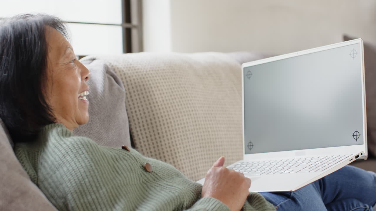 Senior Asian woman video chatting on laptop at home, smiling and waving happily, copy space