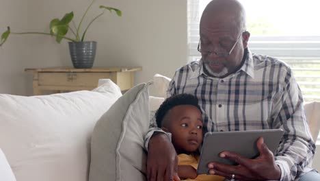 african american grandfather with grandson using tablet on couch at home, slow motion