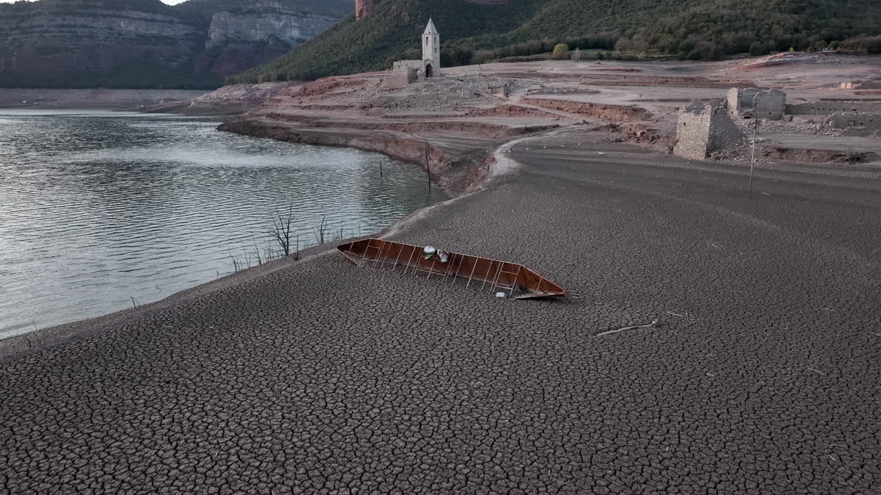 Relitto di un'imbarcazione sulle sponde aride della palude di Sau con rovine di una chiesa sullo sfondo, Catalogna, Spagna.