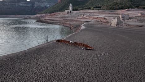 Shipwreck-of-boat-on-dry-shores-of-Sau-marsh-with-church-ruins-in-background,-Catalonia,-Spain