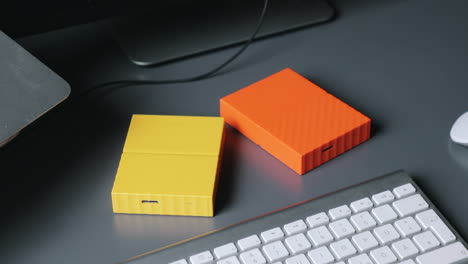 Male hands setting two yellow and orange coloured hard drives on a grey table midst keyboard and mouse