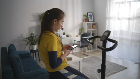 una adolescente está leyendo mensajes y noticias en las redes sociales por celular sentada en una bicicleta estacionaria en casa deporte entrenamiento para la salud y la adicción a internet