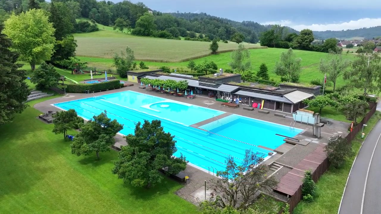 透明な水と豊かな緑に囲まれたスイスのスイミングプールをドローンが旋回上昇する空撮。