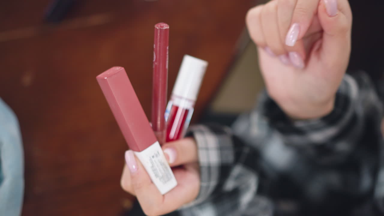 vista parcial de primer plano de una mujer mostrando y analizando múltiples productos cosméticos en la mano, los artículos de horneado, incluidos el lápiz labial y el brillo, se muestran prominentemente en foco con un fondo borroso