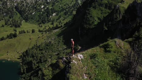 Un-Hombre-Joven,-En-Forma-Y-Fuerte-Está-Solo-Al-Borde-De-Un-Acantilado,-Controla-Su-Dron-Y-Mira-Hacia-Arriba-Para-Disfrutar-De-La-Vista-Durante-El-Caluroso-Día-Soleado-En-Seebergsee,-Suiza