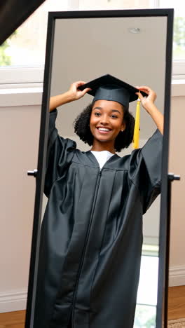 young woman happily trying on graduation cap and gown in front of a mirror