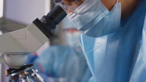 Female-Lab-Worker-Wearing-PPE-Analysing-Samples-With-Microscope-Holding-Test-Tube-Labelled-Covid-19