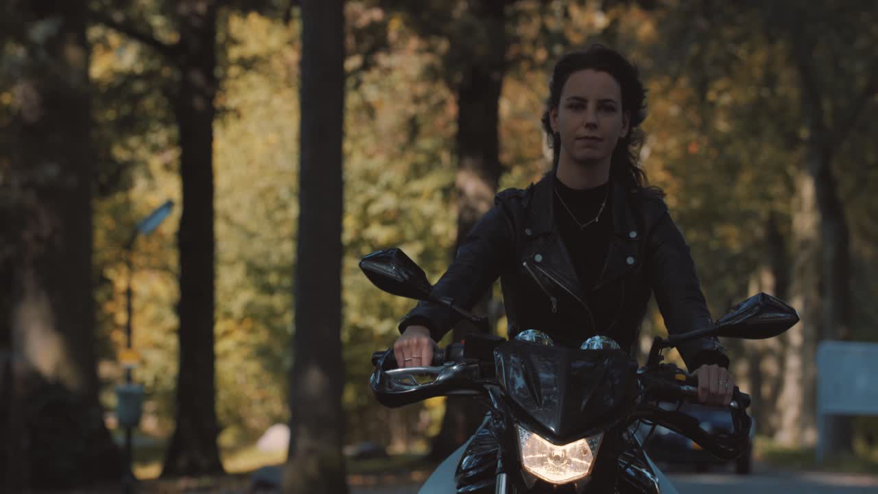 mujer joven europea muy sonriente conduciendo una moto con chaqueta de cuero en el bosque con hojas de otoño doradas vibrantes y coloridas en el día soleado-7