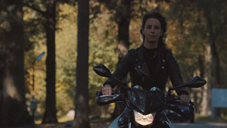 pretty smiling european young woman driving a motorbike wearing leather jacket in forest with vibrant, colorful golden autumn leaves on sunny day-7