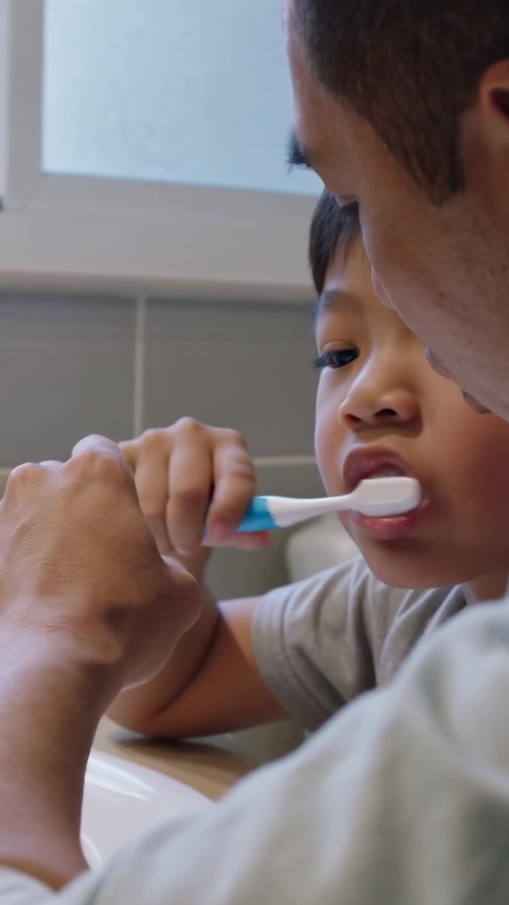 Vertical video: Glancing at tablet, dad light shirt teaching child brushing with blue brush at sink