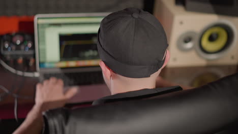 back shot of a man wearing a black cap and shirt, seated on a black chair, working on a computer. a focused work environment, possibly in a recording studio, with audio equipment visible nearby