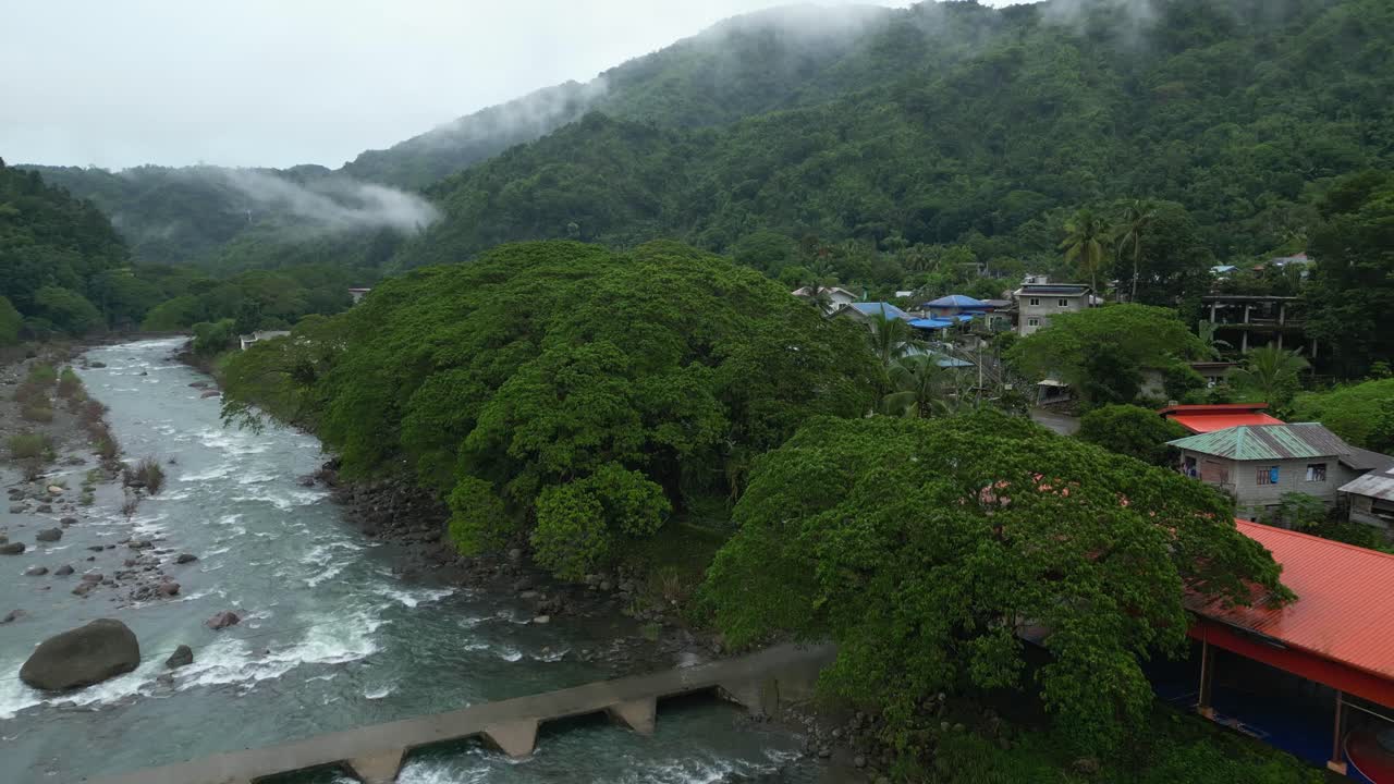 A wide aerial shot hovers above a misty Philippine riverbank. Colorful buildings line the edge of dense forest as rocky rapids flow beneath a narrow bridge