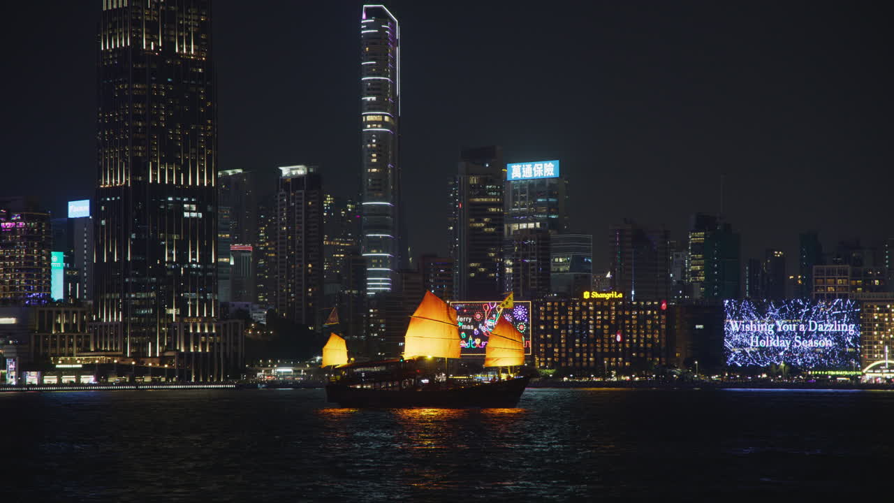 el puerto victoriano de hong kong es un barco de vela rojo tradicional, vista nocturna de los rascacielos de tsim sha tsui con luces de la ciudad.