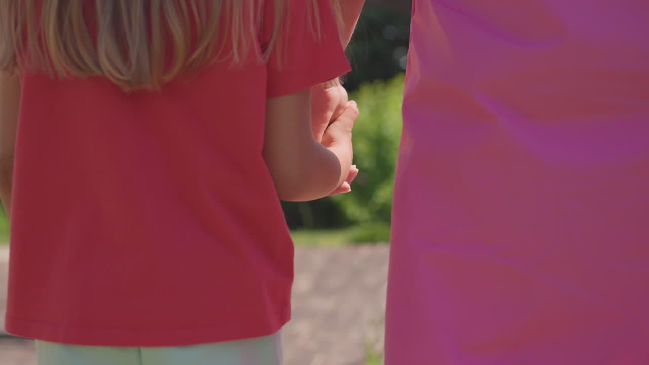 Mother Gently Leads Child Along Bright Garden Walkway In Peaceful Neighborhood, White Mother Wearing Pink Dress Helps Her Blondehaired Child Walk Through Sunny Peaceful Garden Area