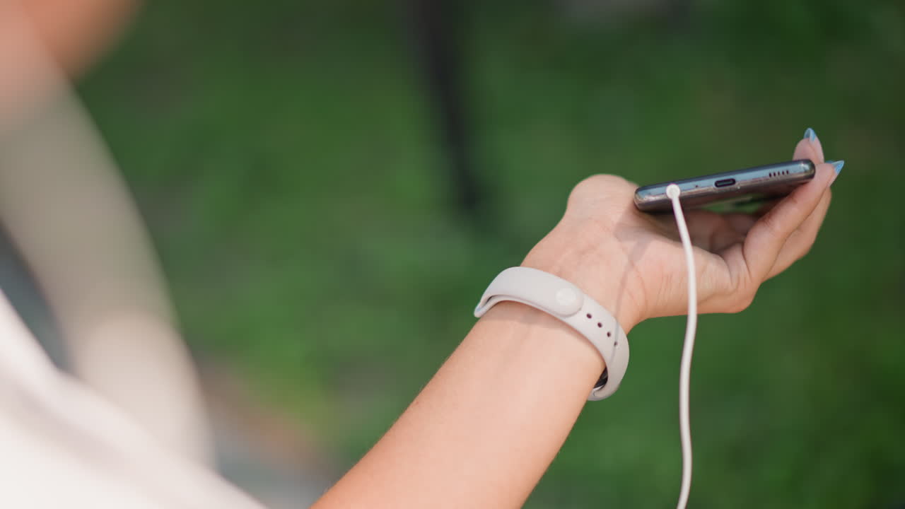 closeup hand adjusting watch near smartphone, fitness enthusiast checks time and notification before run, white strap visible, earbuds cable draped, park path blurred, focused fingers and motion