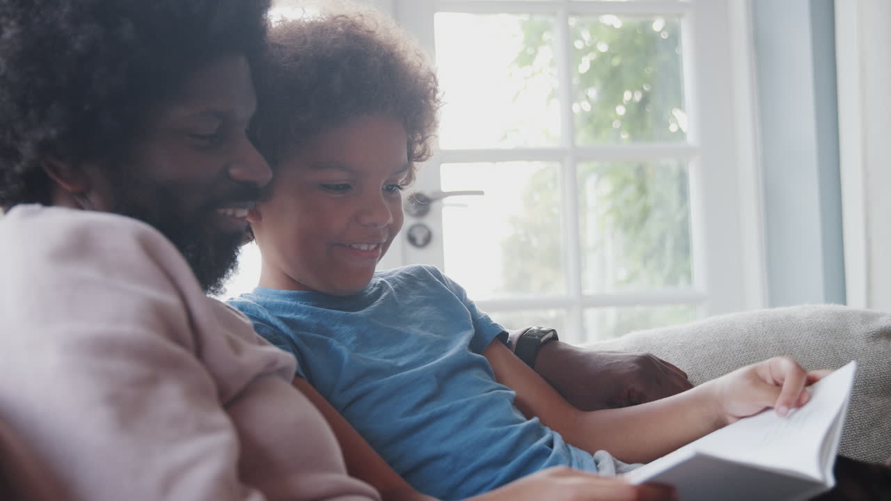 Side view close up of mixed race pre teen boy and his father sitting on a sofa at home and reading a book together, backlit