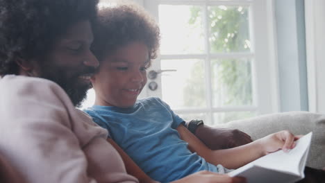 Side-view-close-up-of-mixed-race-pre-teen-boy-and-his-father-sitting-on-a-sofa-at-home-and-reading-a-book-together,-backlit