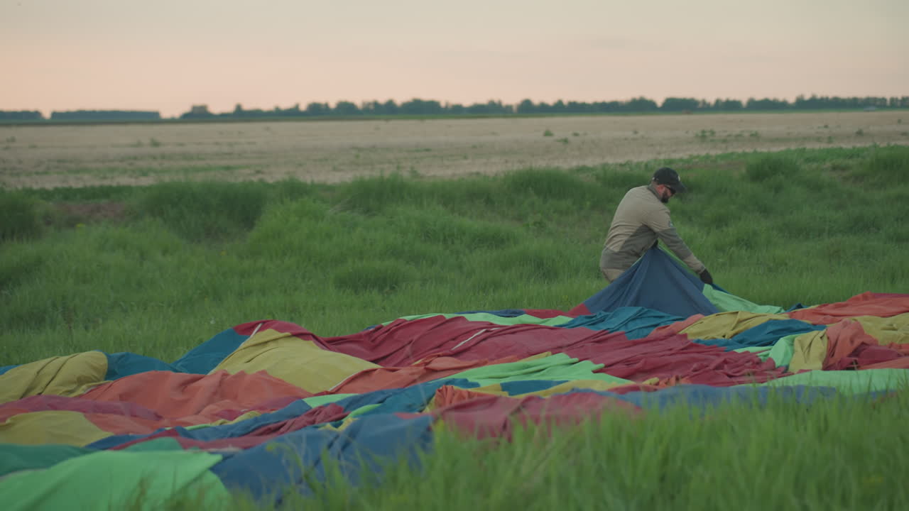 man in cap and gloves sets out colorful hot air balloon canopy across grassy field at dusk under pastel sky during preflight setup in expansive rural landscape