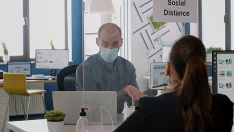 Two-collegues-with-face-masks-working-at-marketing-project-while-sitting-in-company-office