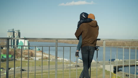 a father carrying his son stands by an iron railing, both watching cars pass by, the father crosses his legs casually, with a blur of the river and cityscape