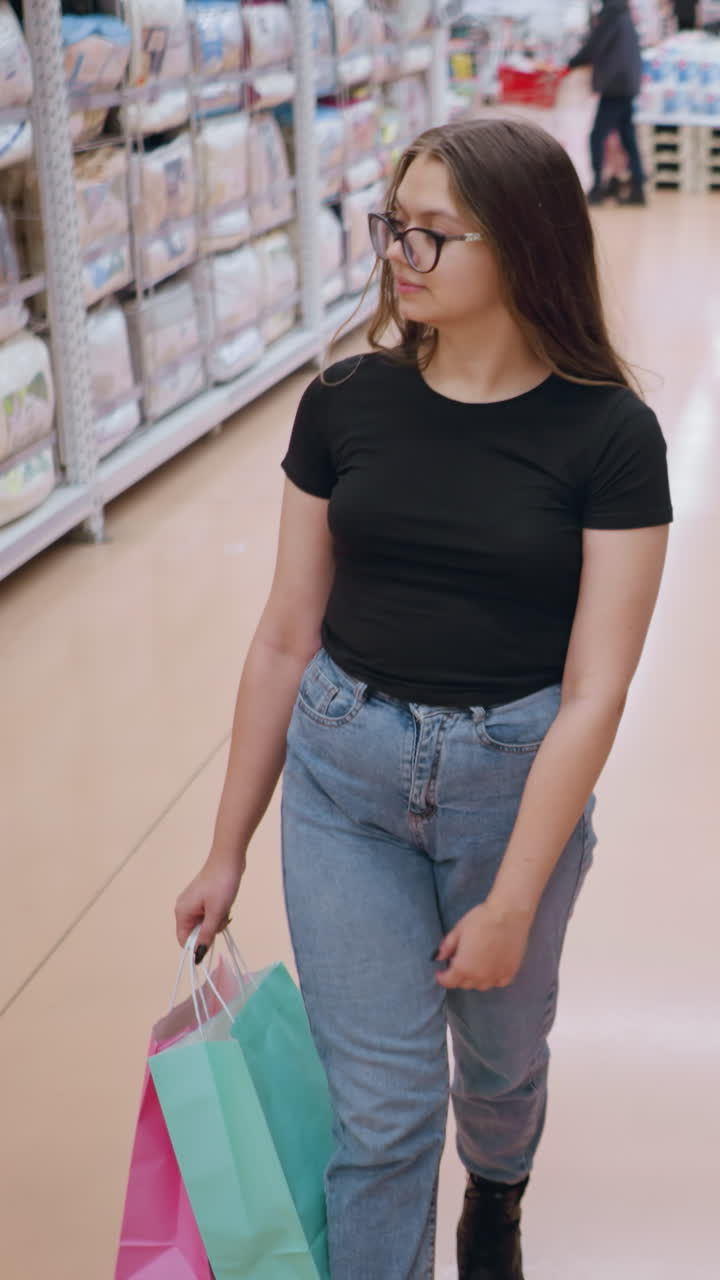 mujer joven con camiseta negra y gafas caminando por el pasillo de la tienda, navegando por las mercancías en los estantes mientras sostiene bolsas de compras, entorno minorista con estantes bien abastecidos y iluminación brillante en el fondo