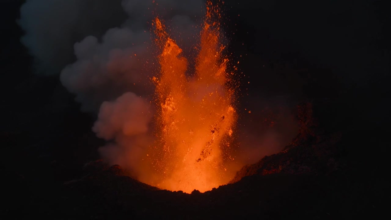Con un'esplosione iniziale di scintille, una bocca del cratere in eruzione espelle piroclasti incandescenti di notte, con una colonna di cenere.