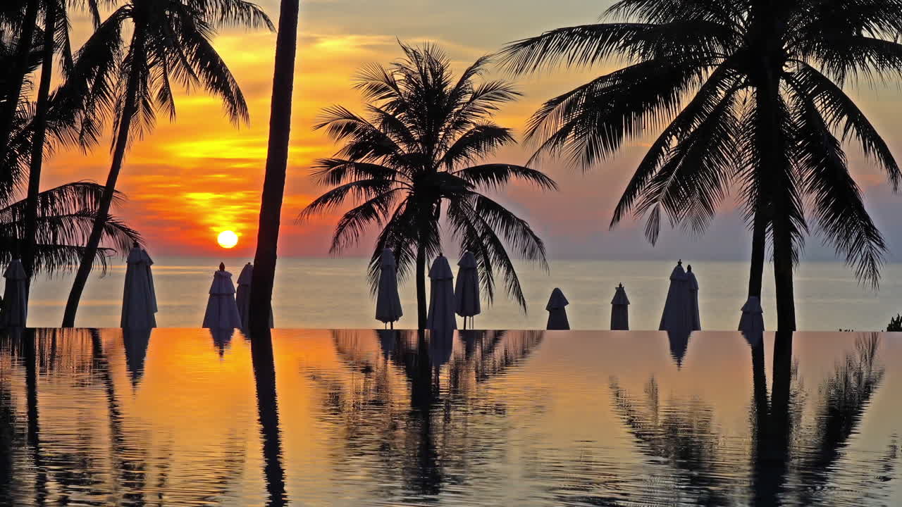 Static shot of an infinity pool during sunset, showing the orange colored sky and a silhouette of palm trees, beach umbrellas and the ocean in the background