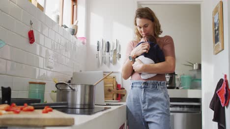 video of caucasian mother with newborn baby in baby carrier cooking in kitchen